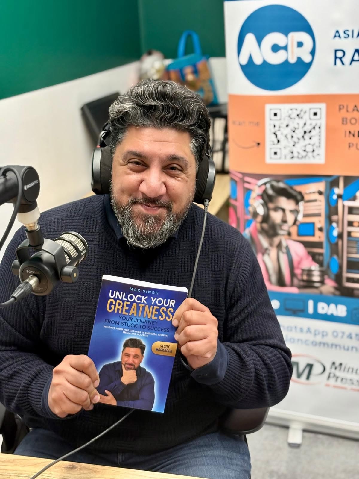Mak Singh at the microphone in the Asian Community Radio MK studio, holding the Unlock Your Greatness study workbook